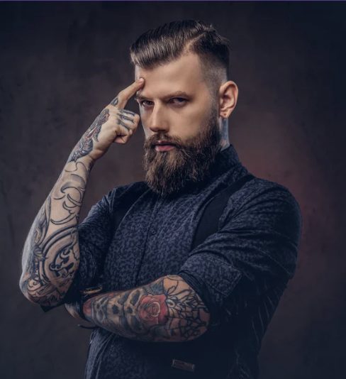 Homem barbudo e tatuado em pose pensativa, em fundo escuro